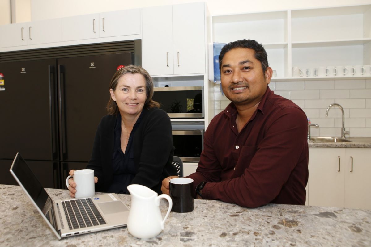 Man and woman in work attire sitting at kitchen island, laptop on island top, kitchen cupboards, microwave and fridge in background.
